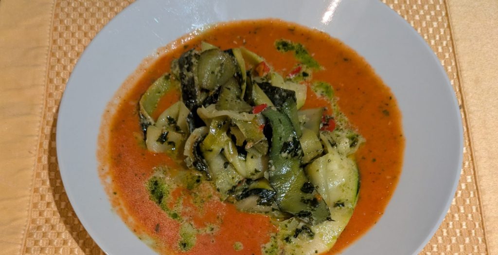 Roasted tomato soup with zucchini ribbons on top served in a white bowl over a golden placemat.
